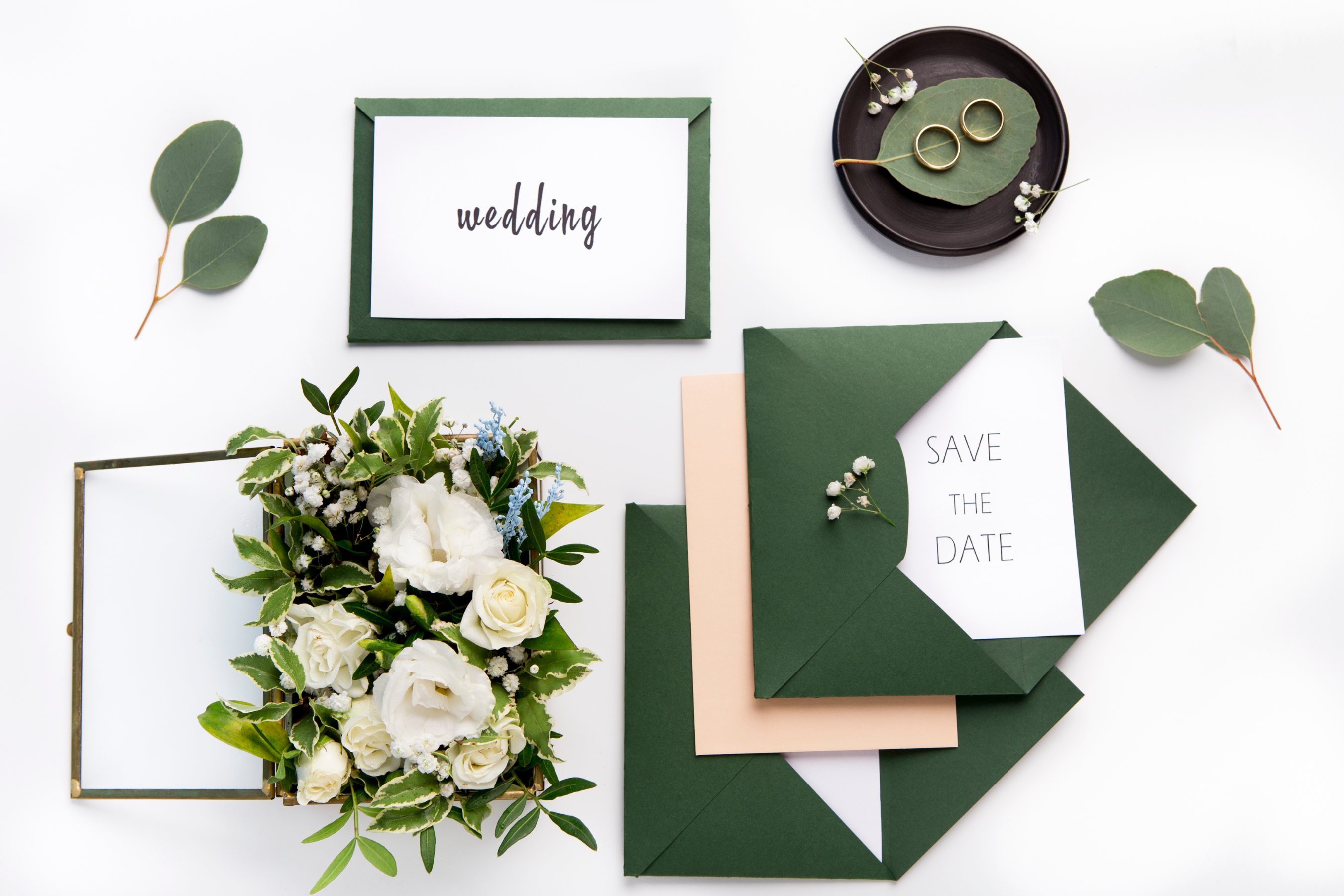Green and white wedding save the date cards with envelopes, floral arrangement, and wedding rings styled in an elegant flat lay.