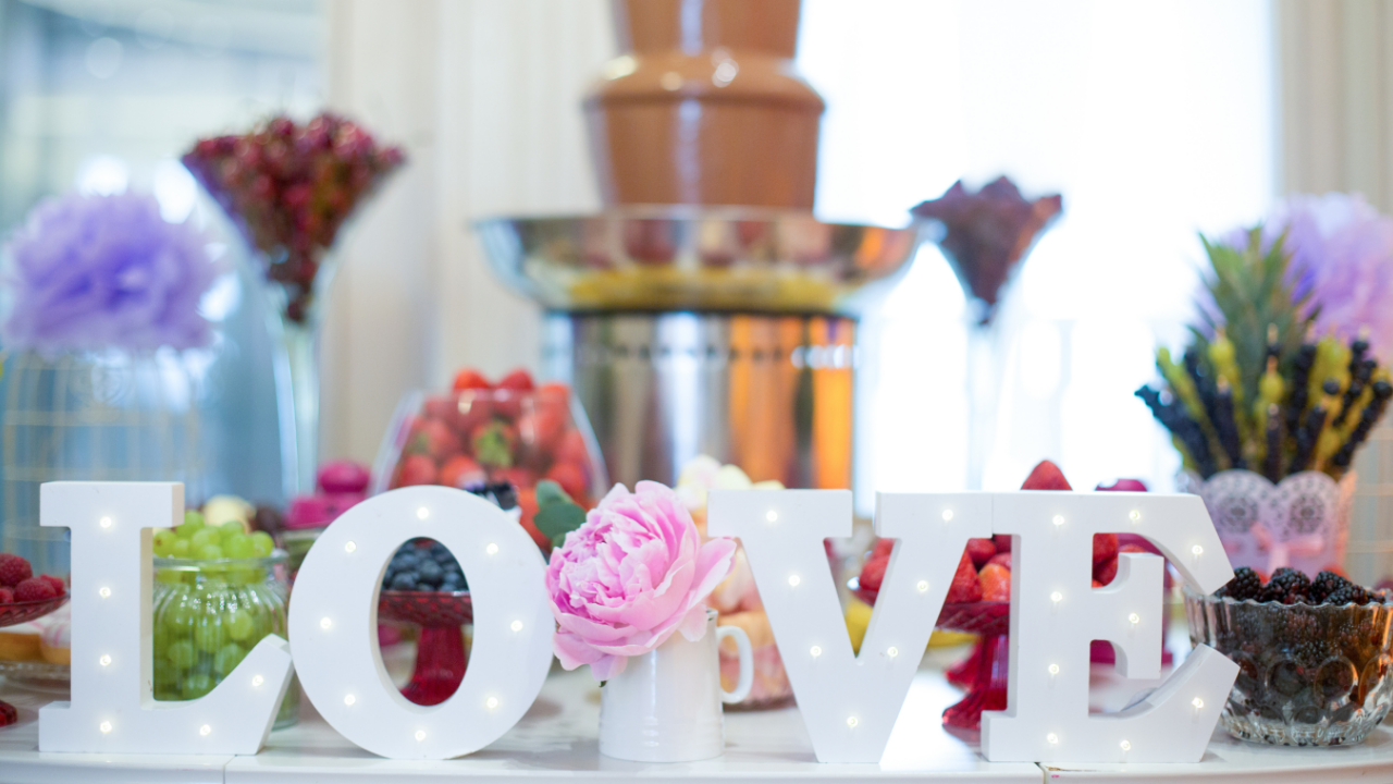 Elegant LDS wedding reception dessert table featuring a chocolate fountain, colorful fruit displays, and illuminated LOVE letters surrounded by flowers and sweet treats.