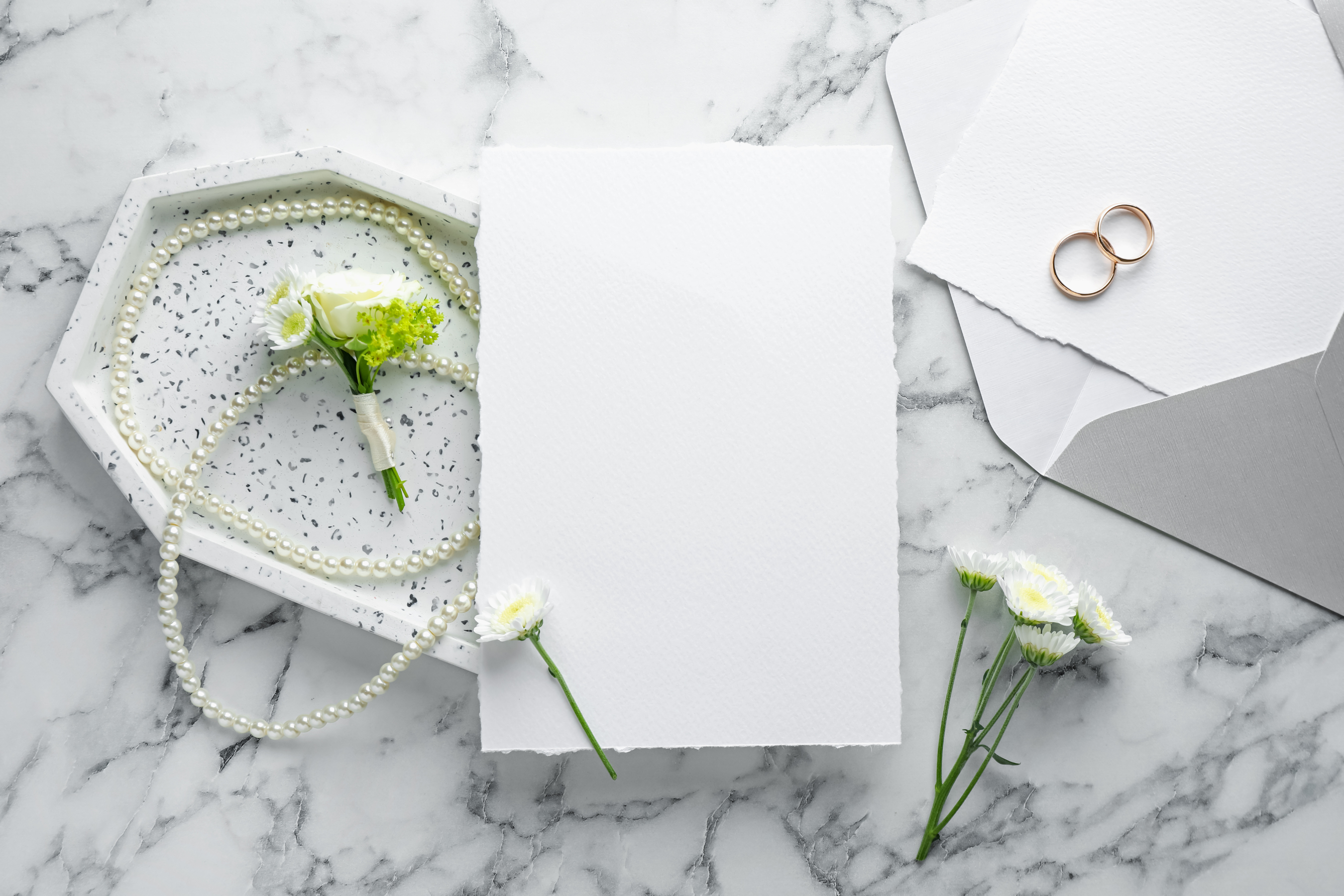 Minimalist DIY wedding invitation setup with blank textured card, envelope, boutonniere, pearls, and wedding rings arranged on a marble surface.