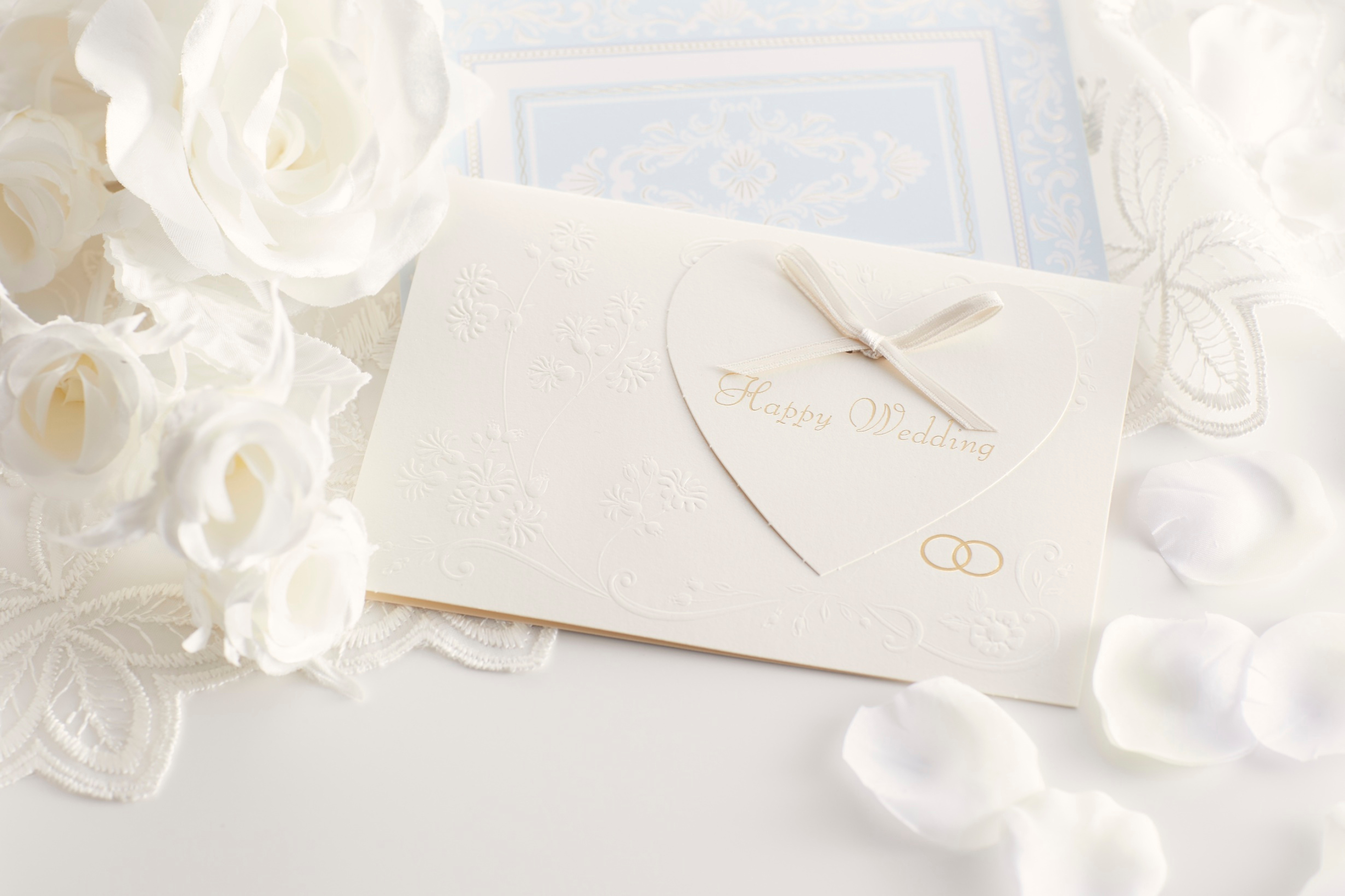 Elegant ivory wedding invitation with embossed floral details, heart-shaped card, and ribbon, surrounded by soft white flowers and lace accents.