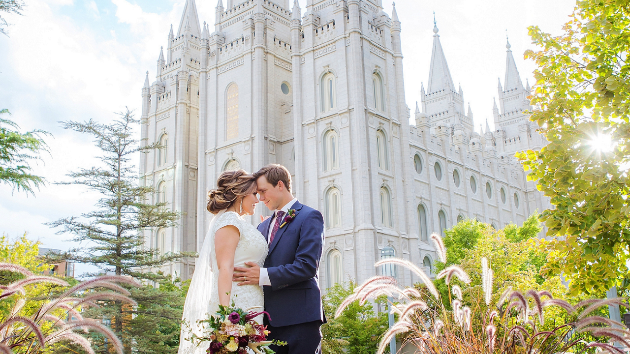 Bride and groom embracing outside the Salt Lake Temple after their temple sealing, surrounded by gardens and sunlight on their wedding day.
