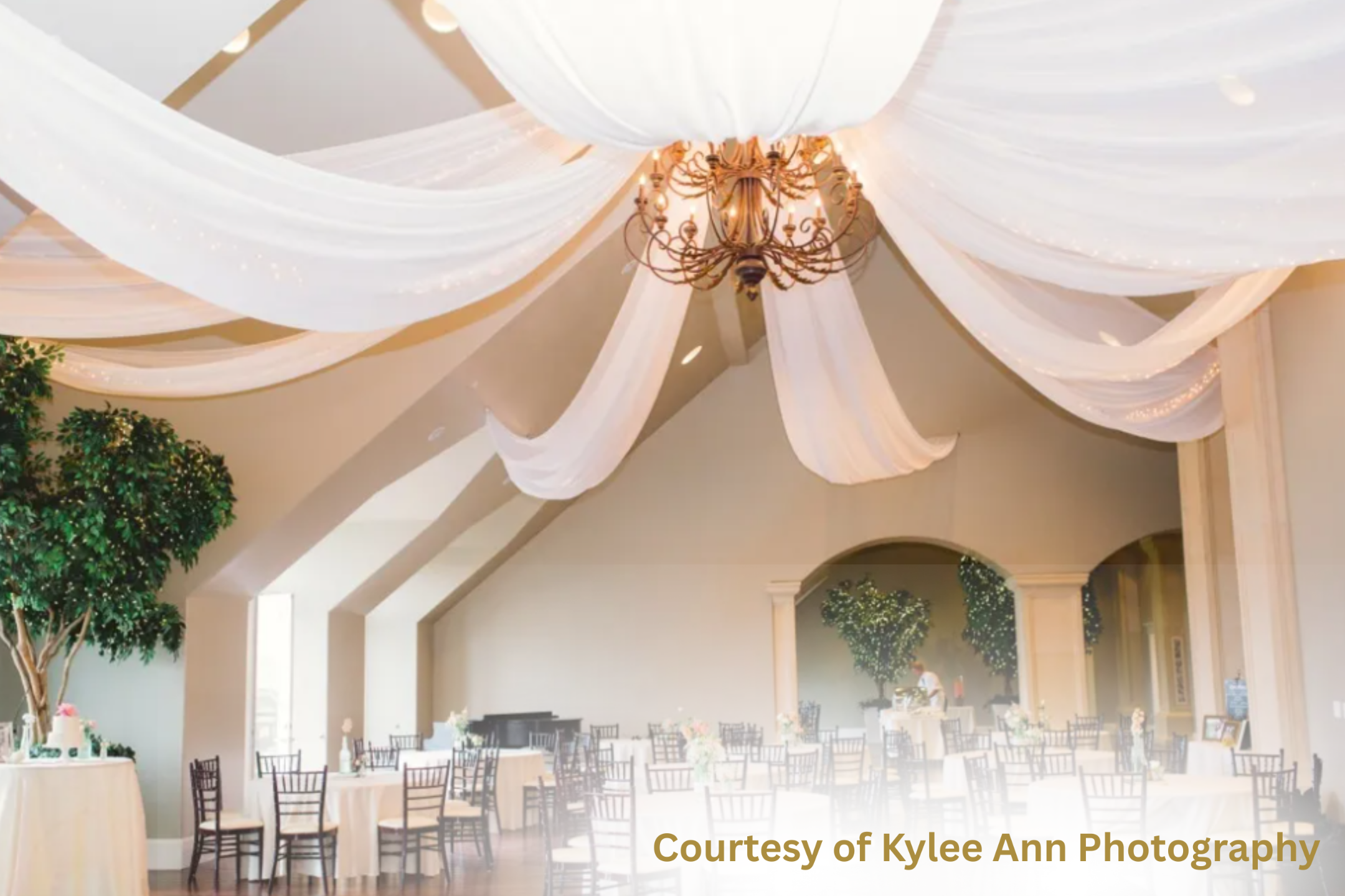 Elegant wedding reception venue featuring flowing white ceiling drapery, a bronze chandelier, round guest tables with white linens, and soft floral centerpieces in a spacious ballroom-style setting.