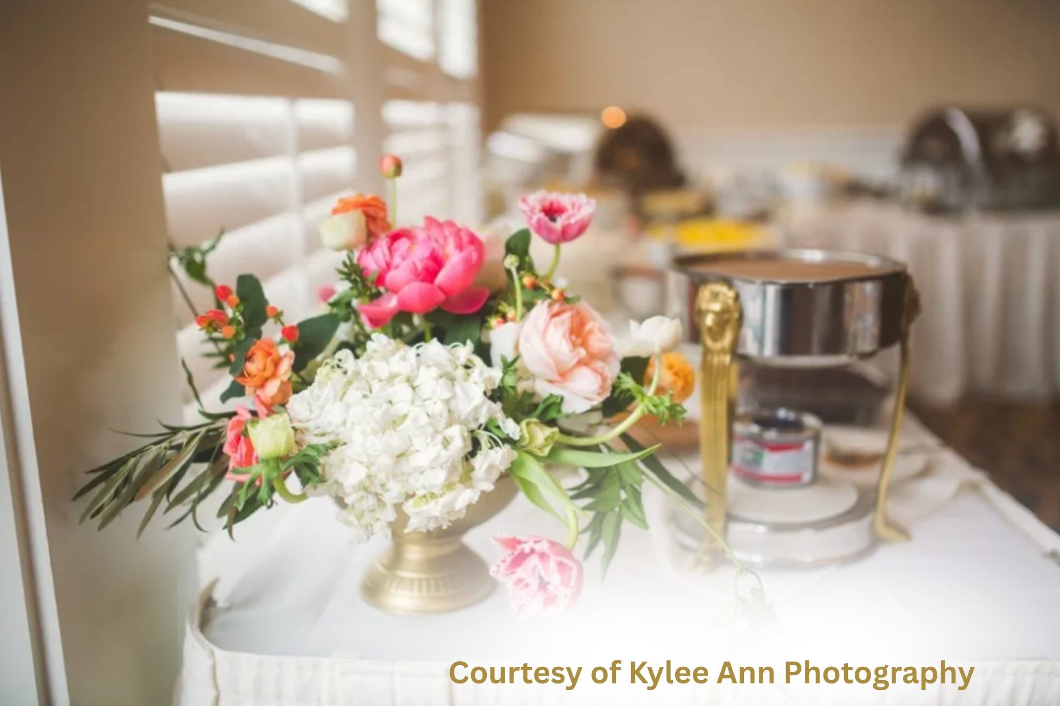 Elegant LDS wedding catering table featuring a gold vase filled with white hydrangeas, coral roses, and bright pink peonies beside a silver chafing dish, softly lit by natural light through white shutters.