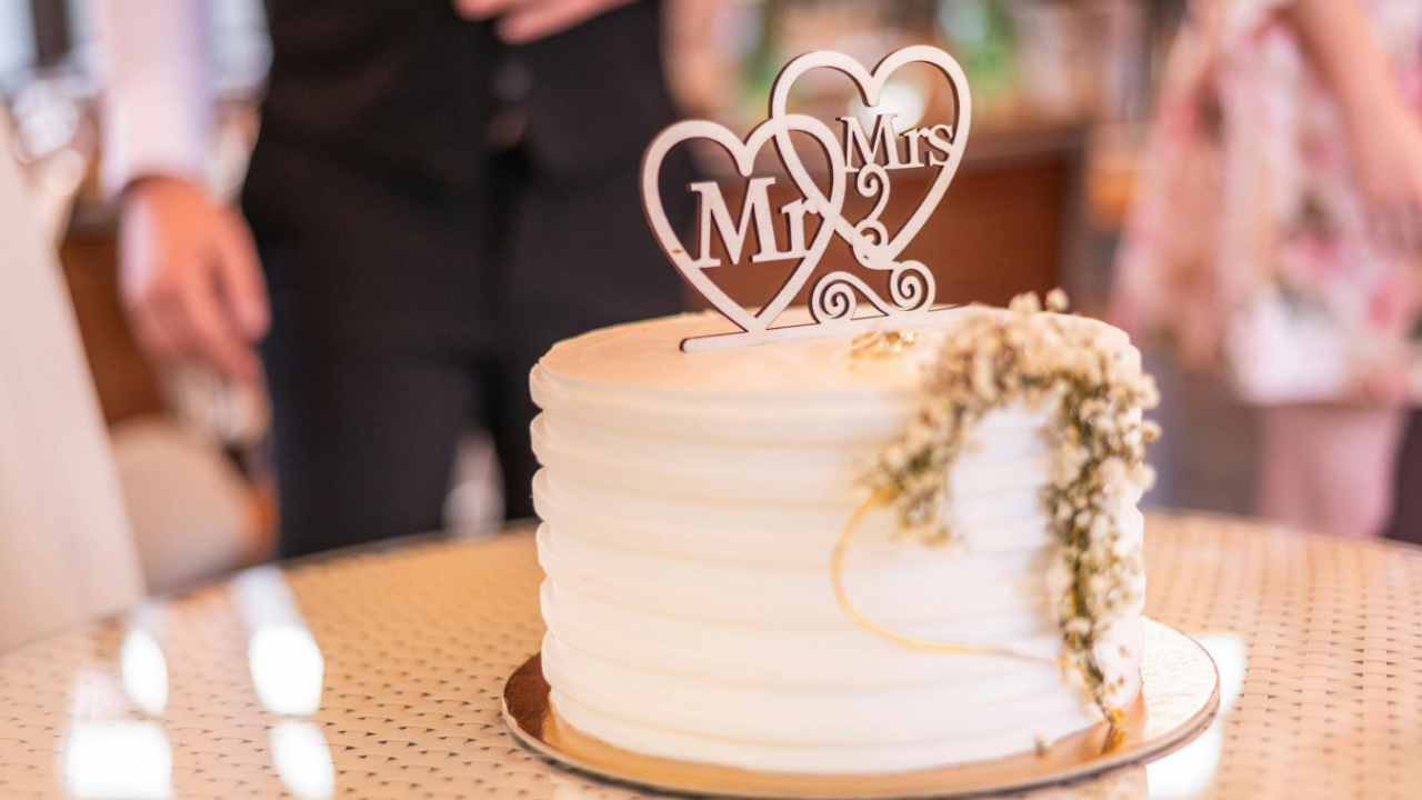 Elegant white wedding cake with textured buttercream frosting, delicate baby's breath floral accents, and a romantic "Mr & Mrs" heart-shaped cake topper, displayed at a wedding reception with the bride and groom softly blurred in the background.