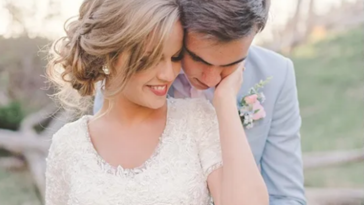 Romantic LDS bride and groom embracing outdoors at sunset, with the bride wearing a modest lace wedding dress and soft updo hairstyle while smiling gently beside her groom in a light gray suit.