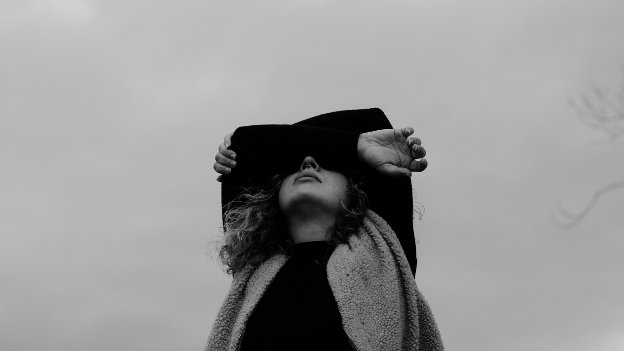 Black and white photo of a woman shielding her face with her arm against a grey sky, carrying the weight of hyper-independence.