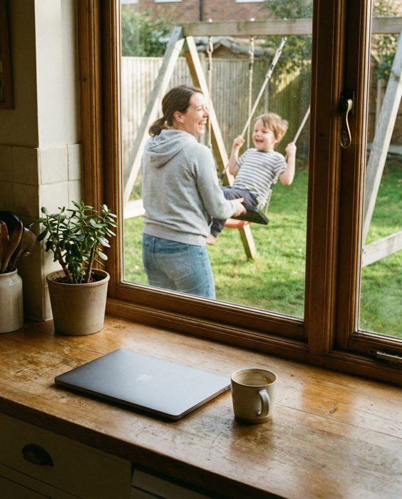 A mum pushing her child on a garden swing, seen from a kitchen window with the laptop closed on the sill