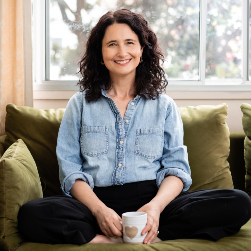 Lea sitting cross-legged on a green sofa, smiling and holding a mug with a heart on it