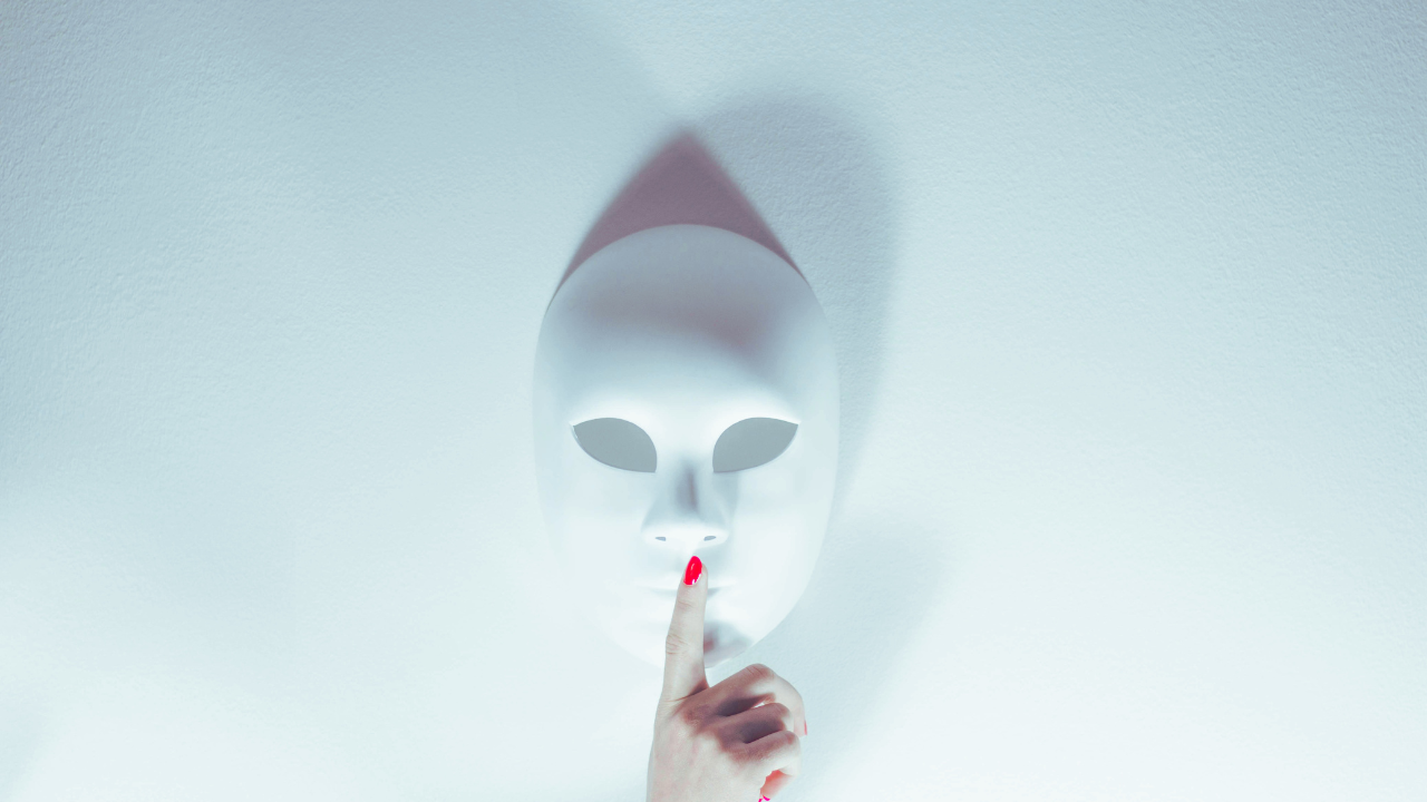 White theatrical mask with a finger held to its lips, symbolizing silence and avoiding difficult conversations.