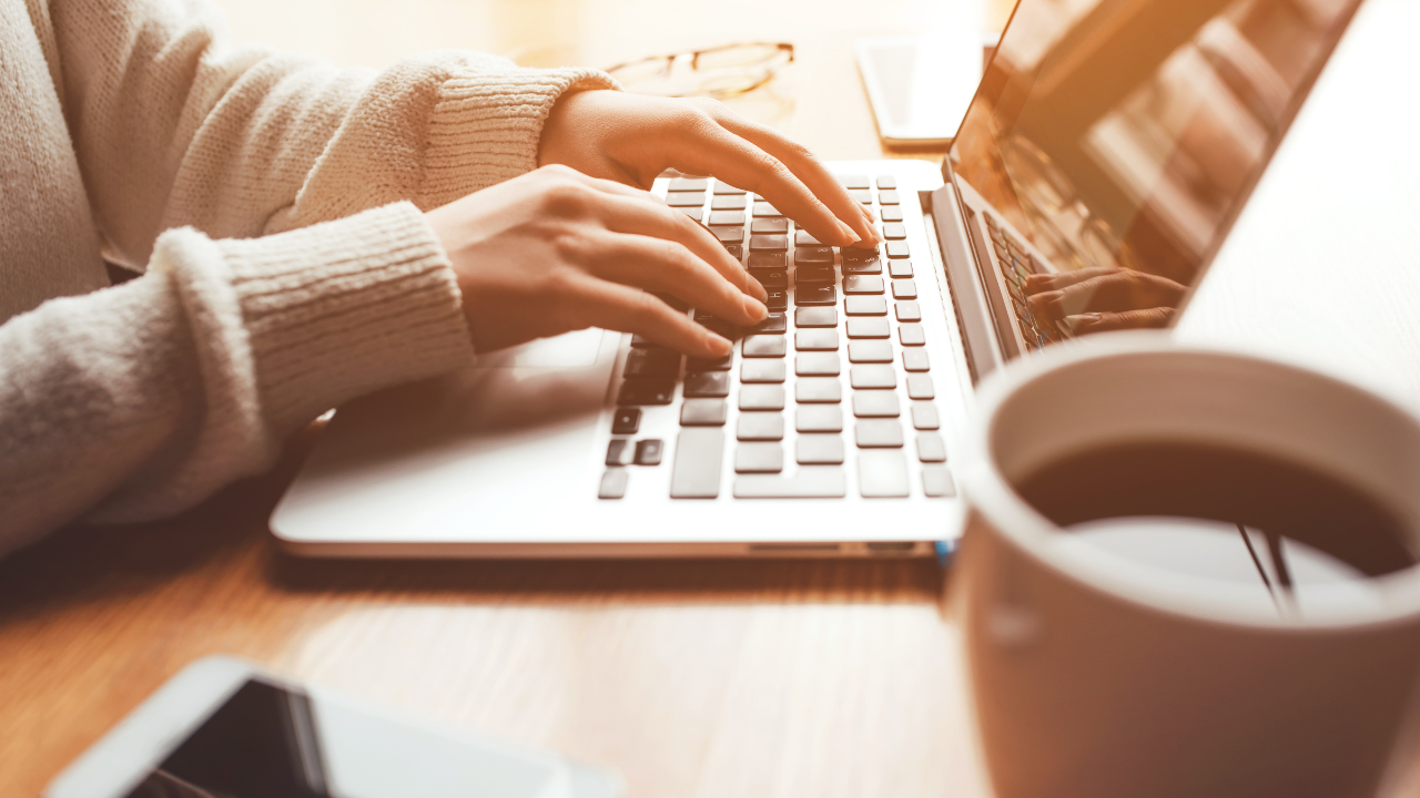 A person typing in their laptop with a coffee on top of their table