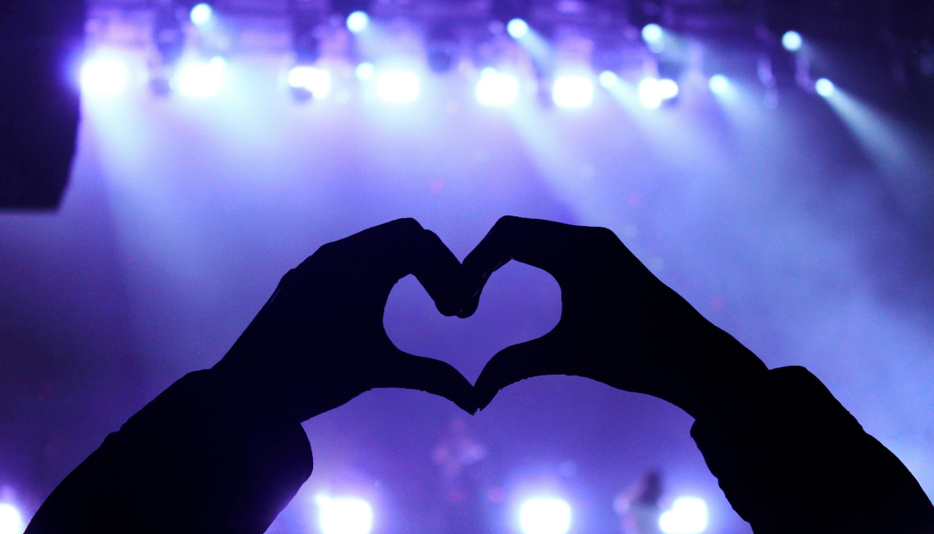 Photo of a fan making a heart with their hands in the crowd during a concert.