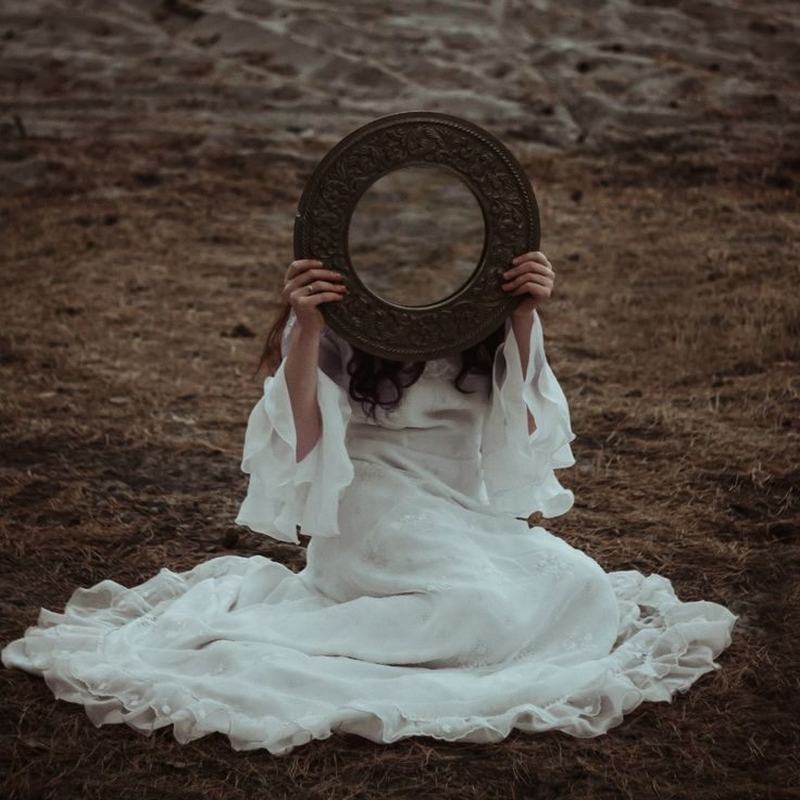 A woman in a white dress sitting on the earth, holding an antique mirror over her face