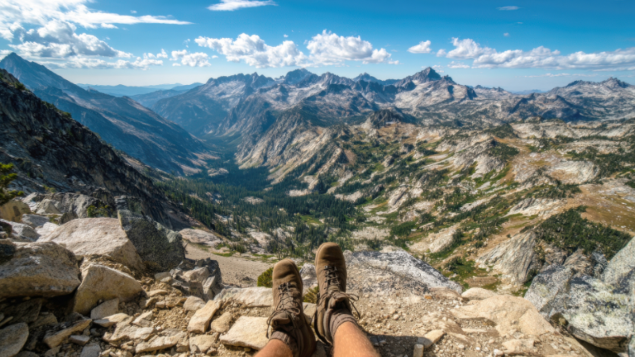 The feet of a person with hiking boots on sitting on the ledge overlooking a breath taking view of rugged mountain range.