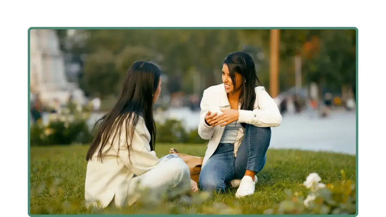 Dos mujeres conversan sentadas en un parque; una habla mientras la otra escucha.