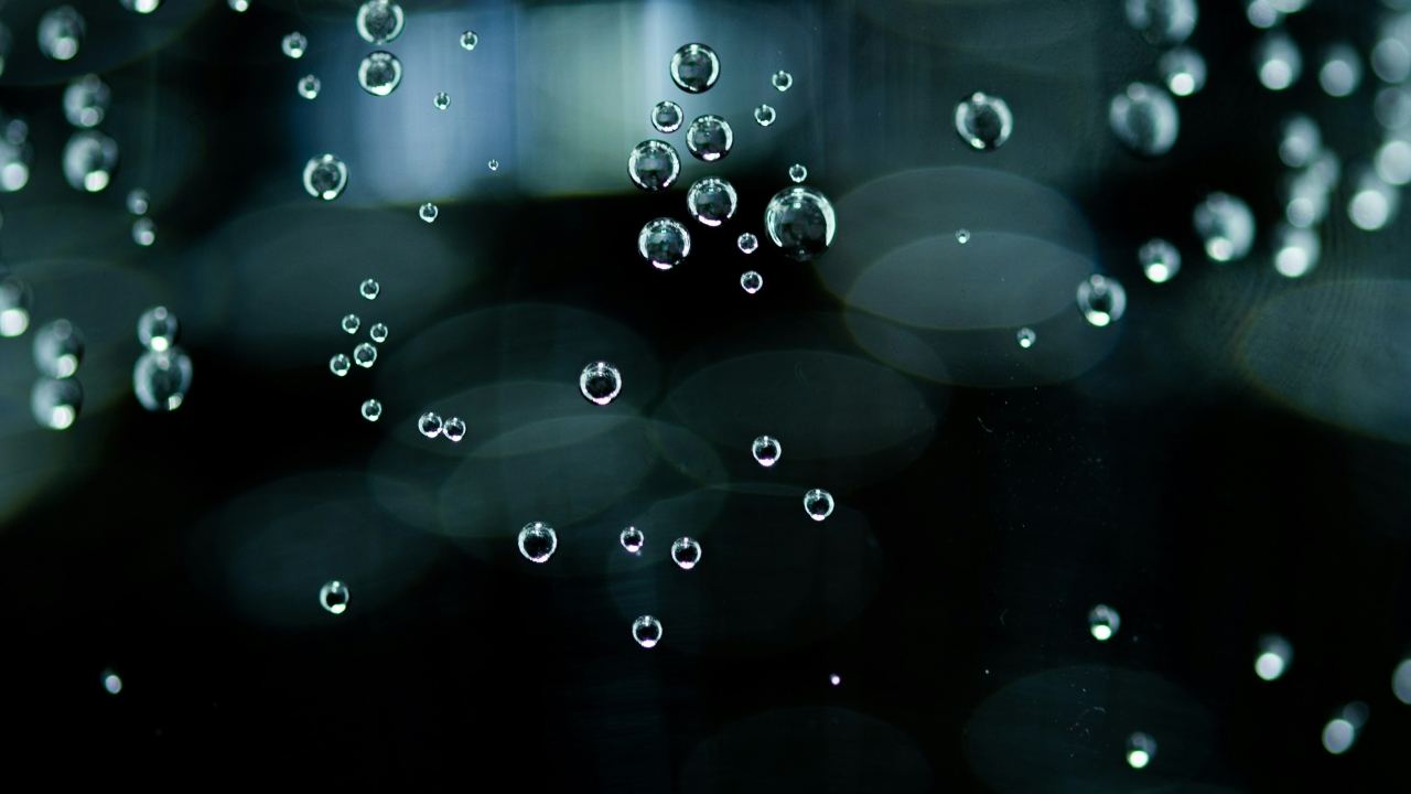 A close-up, macro photo showing numerous tiny, spherical gas bubbles rising through a clear liquid against a dark, moody background.