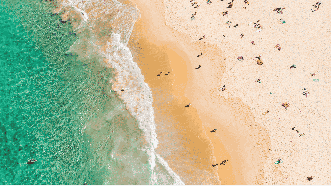 An overhead image of a beach—the green-blue ocean is on the left, white waves crashing in the middle, and tiny “dots” of people scattered on cream sand on the right.
