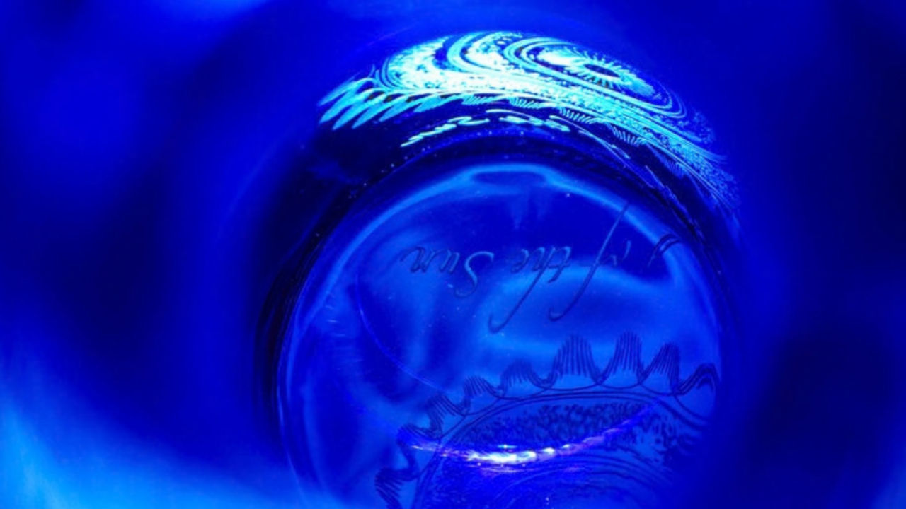 Looking down through the interior of a deep blue cobalt glass bottle. Light reflects off the curved inner walls, creating a soft, luminous cyan swirl. At the bottom, the sandblasted "Song of the Sun" CymaGlyph and elegant cursive script are visible, illuminated by a bright point of light shining through the glass.