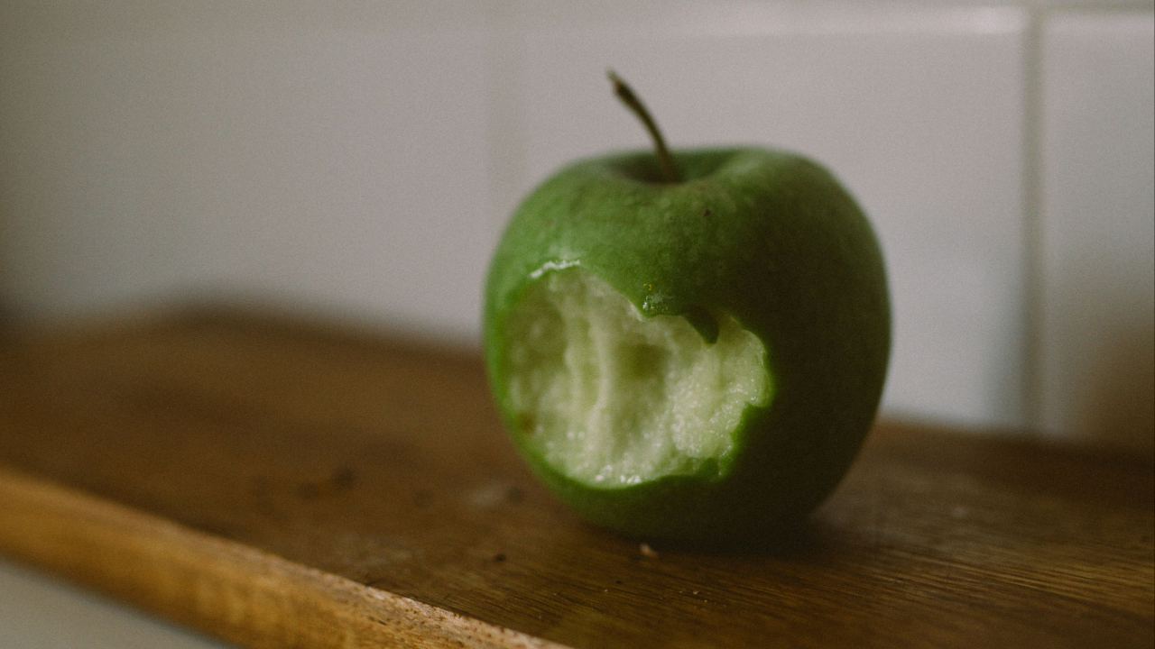 A close-up photograph of a plump, vibrant green apple resting on a rustic wooden table. A single bite mark is visible, revealing the juicy, bright white flesh inside