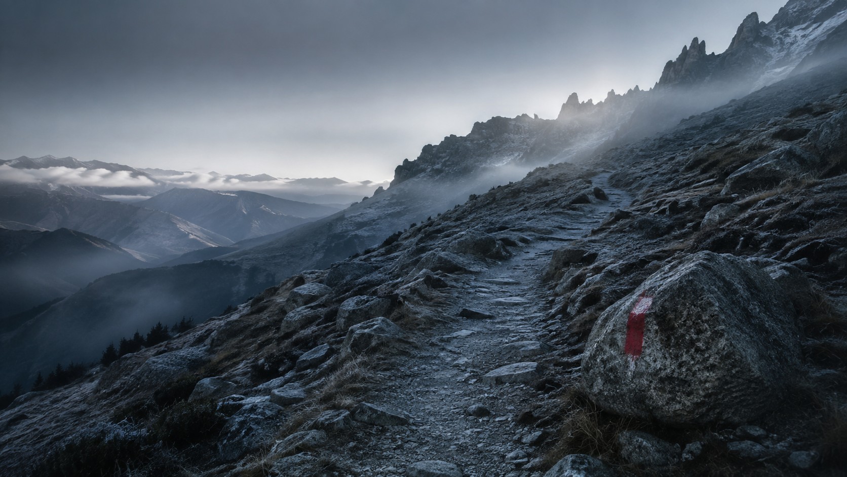 Sentiero di montagna all'alba che sale nella nebbia verso un crinale dalla fine di una strada sterrata; su un masso, un segnavia rosso appena dipinto.