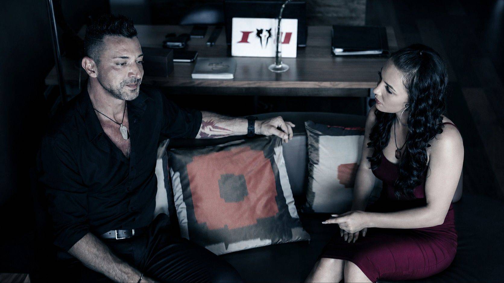 Aivia Demon in camicia nera a colloquio con una donna in abito elegante, seduti in un salotto in penombra.
