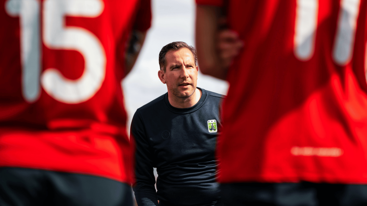 coach standing on a soccer field, framed between blurred players in red jerseys during a team huddle.