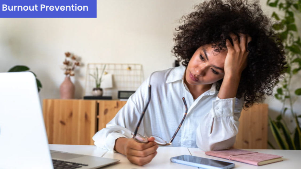 Woman sitting at a home office desk with a laptop, resting her head in one hand and holding glasses in the other, with a tired and overwhelmed expression. A smartphone and notebook are on the desk beside her. Text overlay in the upper left reads "Burnout Prevention" in white on a blue background.