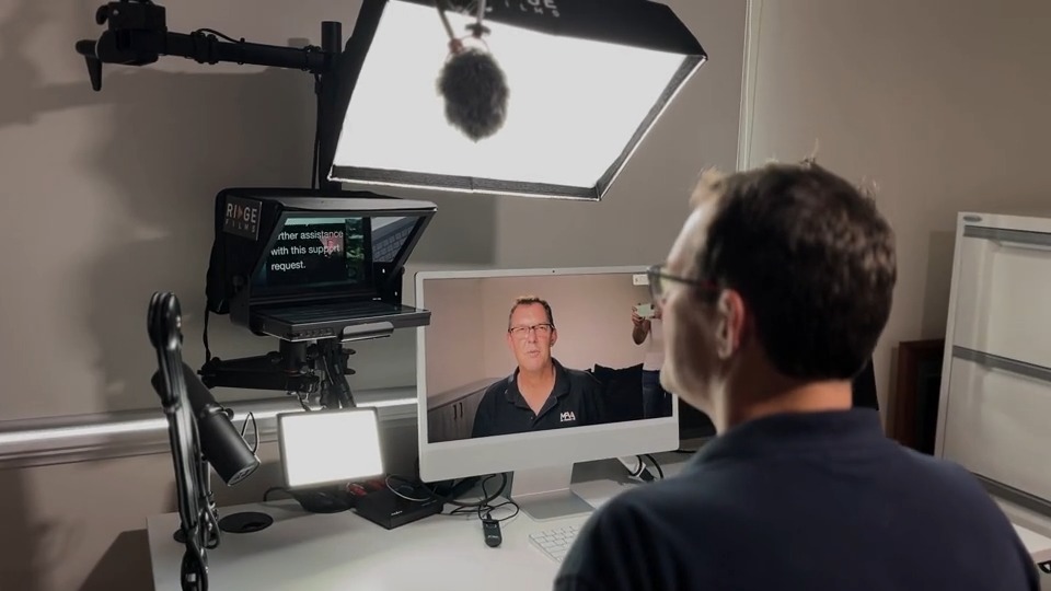 Over-the-shoulder view of a man on a video call at an iMac beside his teleprompter and softbox