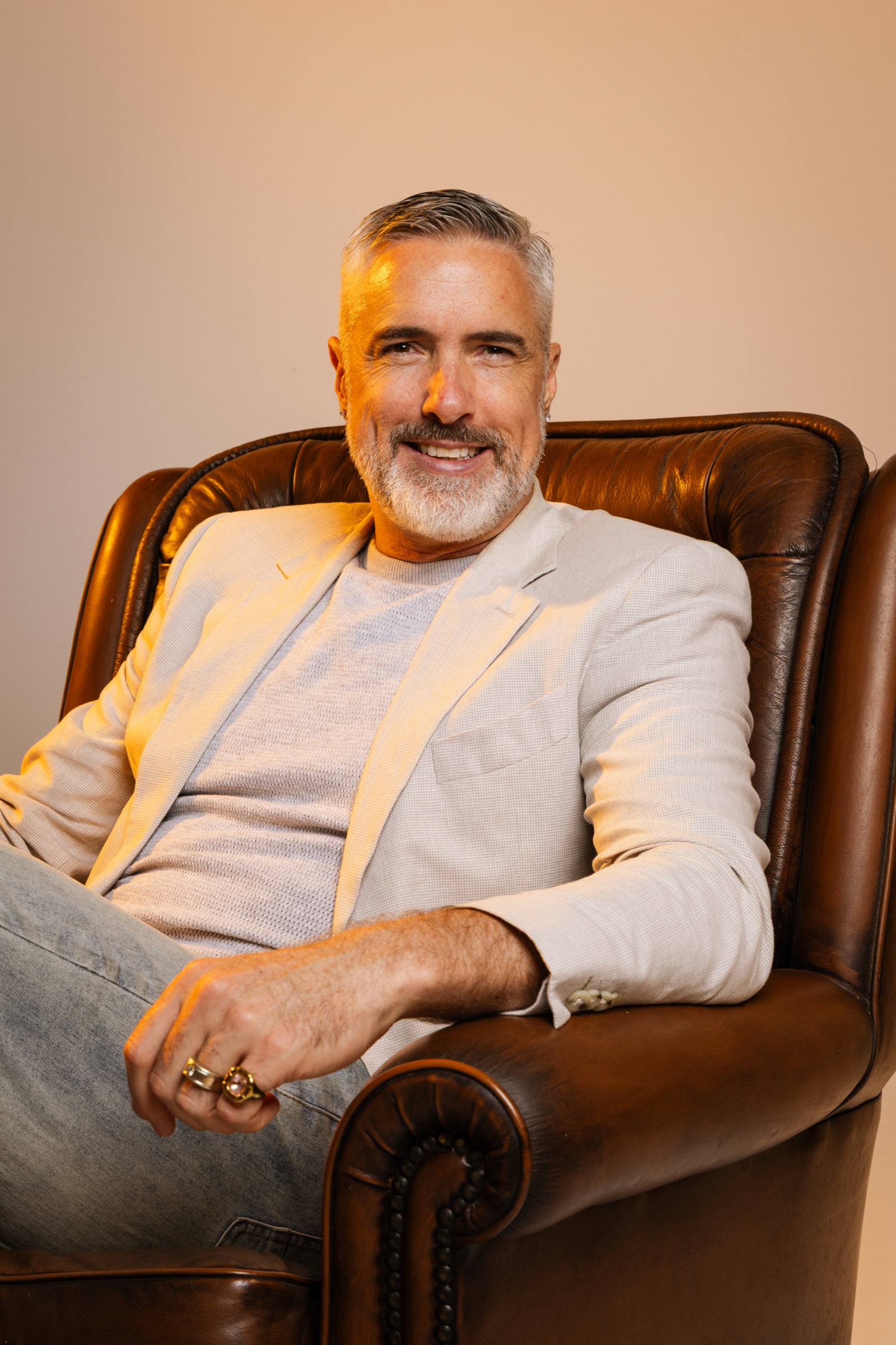 Chris Schwager in a beige blazer, seated in an armchair, smiling warmly at the camera