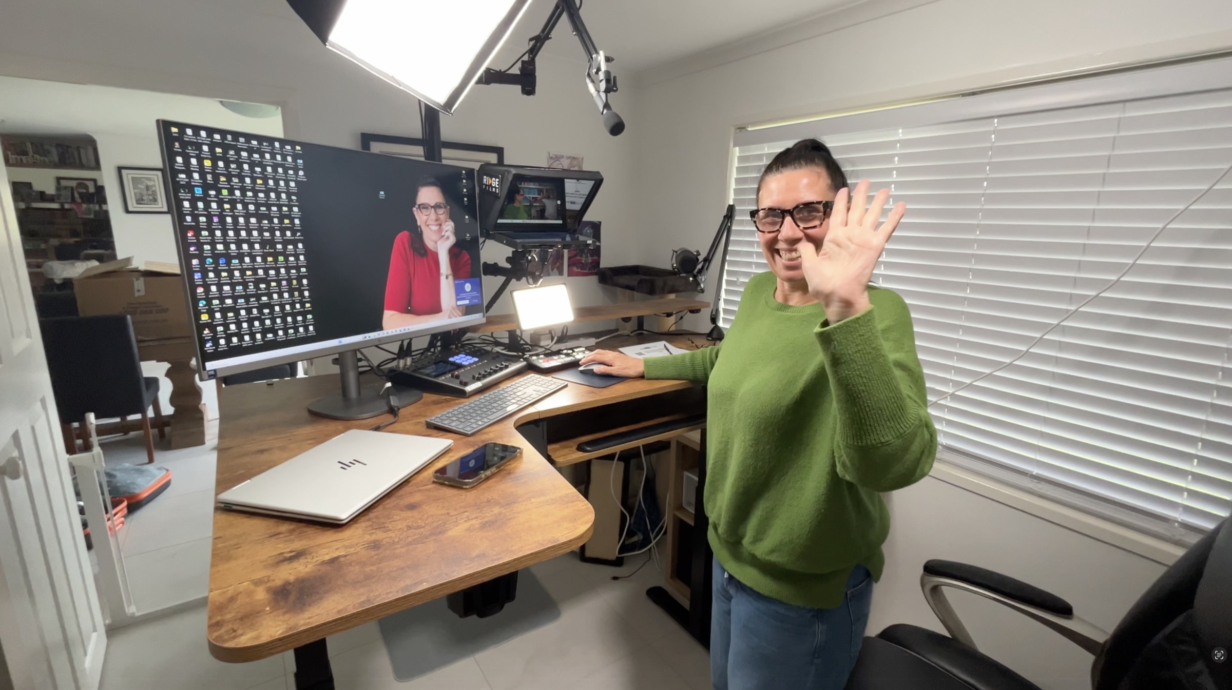 A Ridge client in a green jumper waving beside her desk studio in her home office, softbox and teleprompter set up