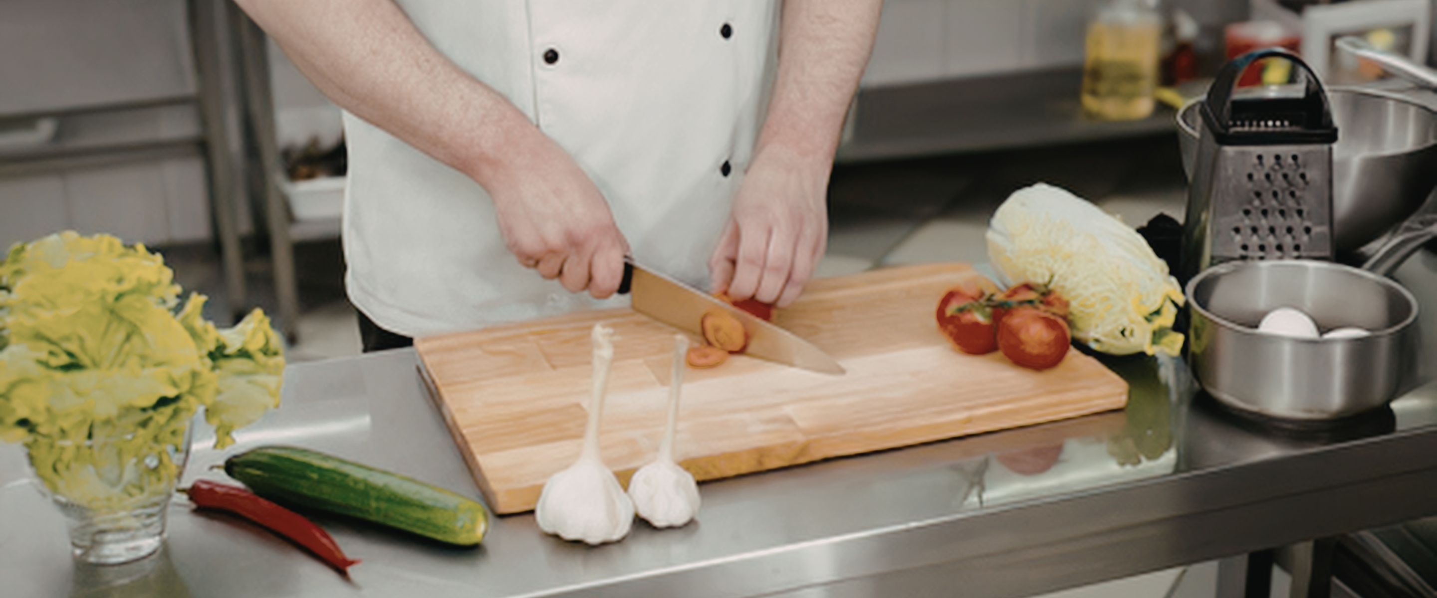 A cook in kitchen whites slices tomatoes on a wooden board in a stainless steel kitchen.