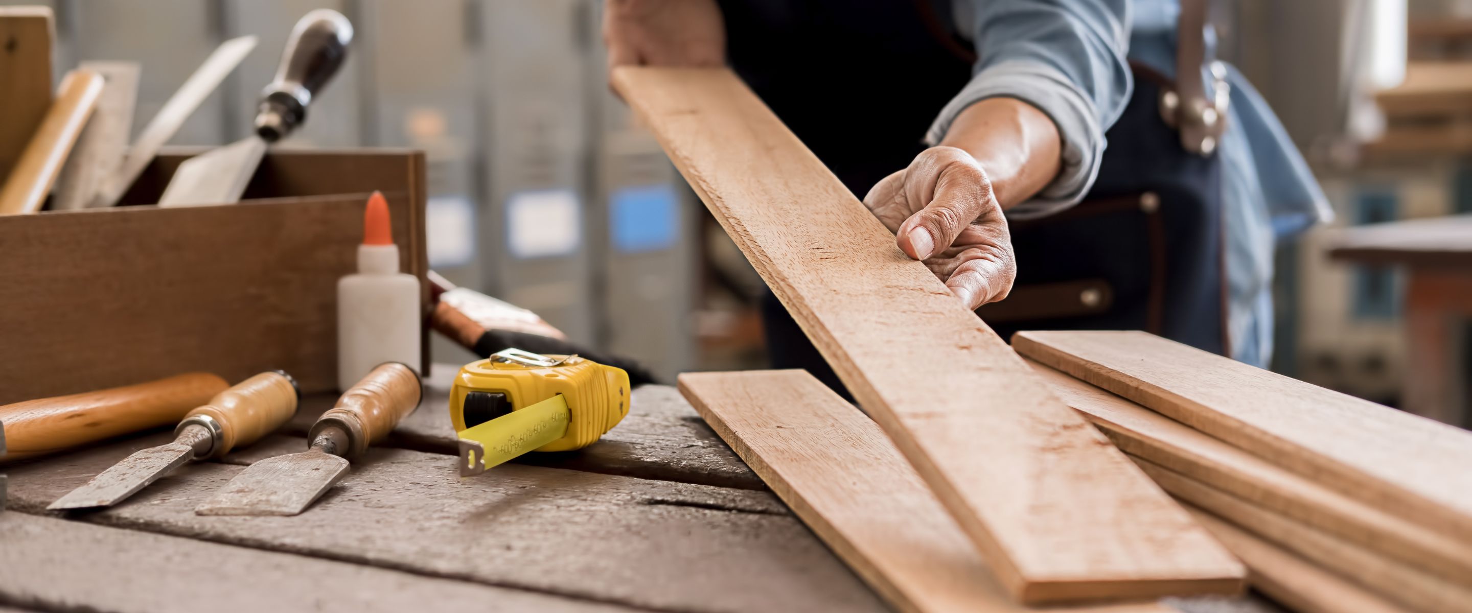Hands working a length of timber at a workbench laid with chisels, a tape measure and glue.