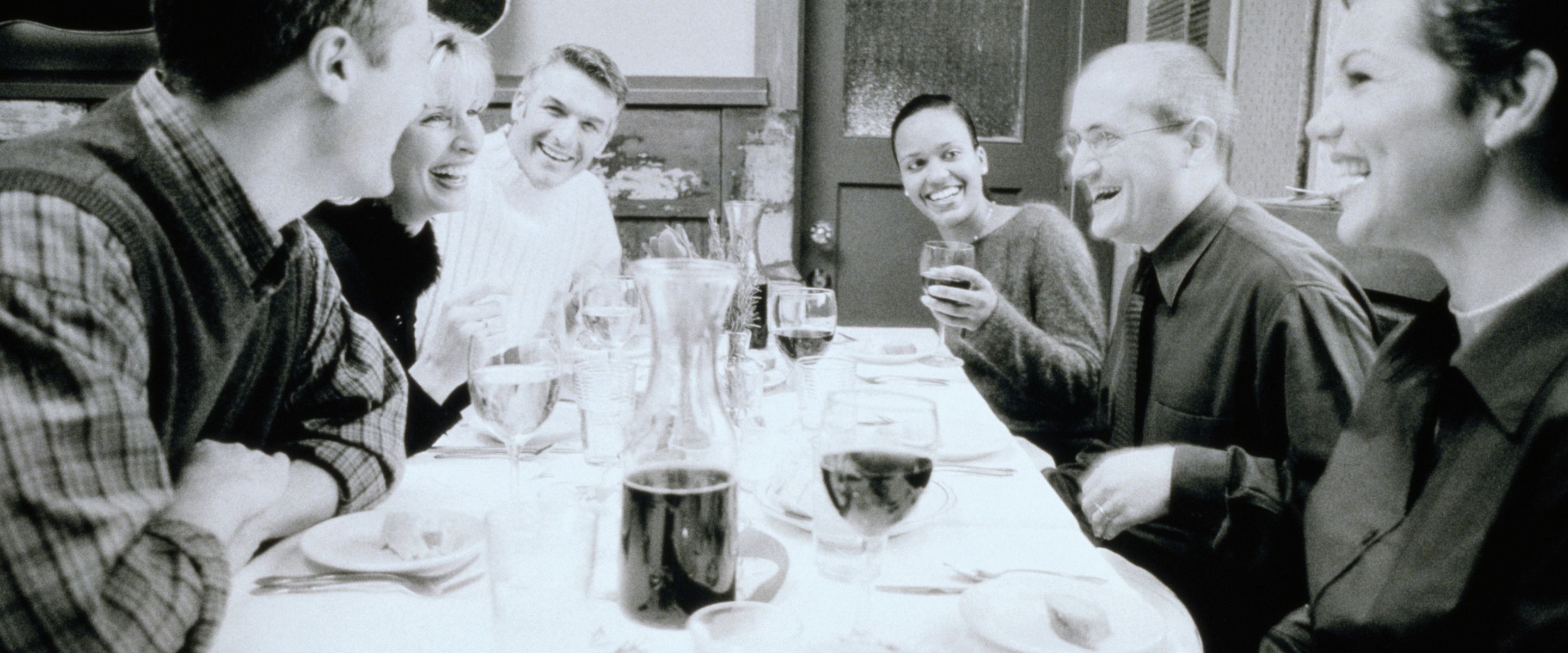 A black and white photograph of six people laughing around a long dinner table set with glasses.