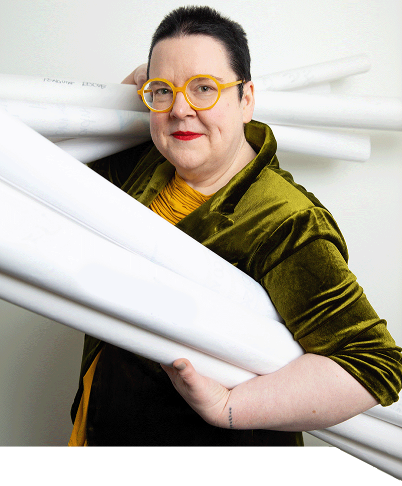 Brandy Agerbeck smiles, looking to camera, wearing deep green glasses, red lipstick, and a bright chartreuse jacket. The the background is a shelf holding Neuland BigOne markers in rainbow order.