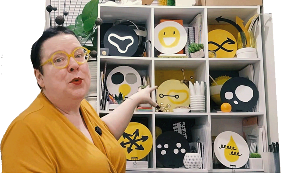 Brandy Agerbeck pointing at her 3x3 bookcase that mirrors the 9-module structure of her comprehensive visual thinking course