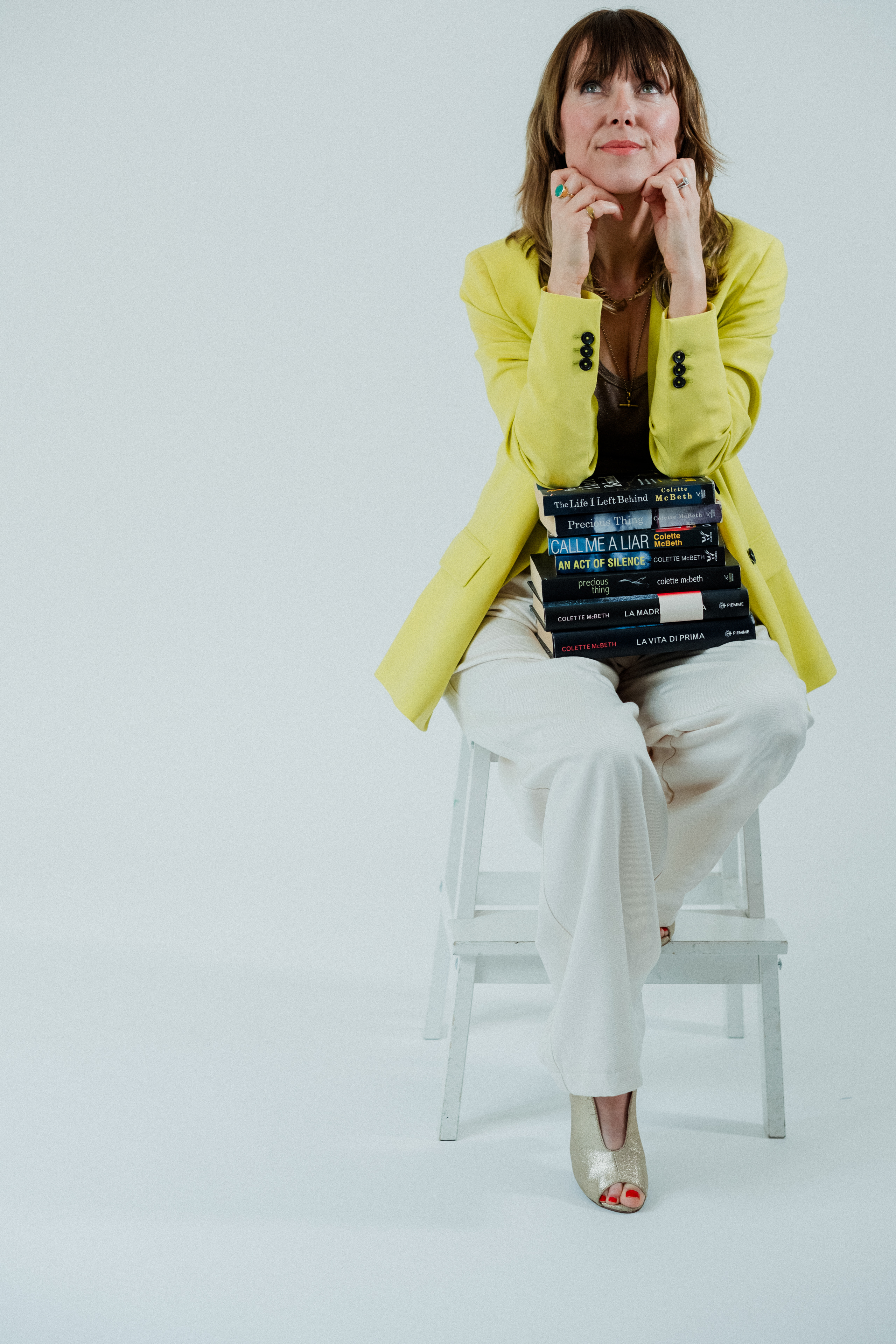 Colette McBeth in a yellow blazer, sitting on a stool with a stack of her novels on her lap