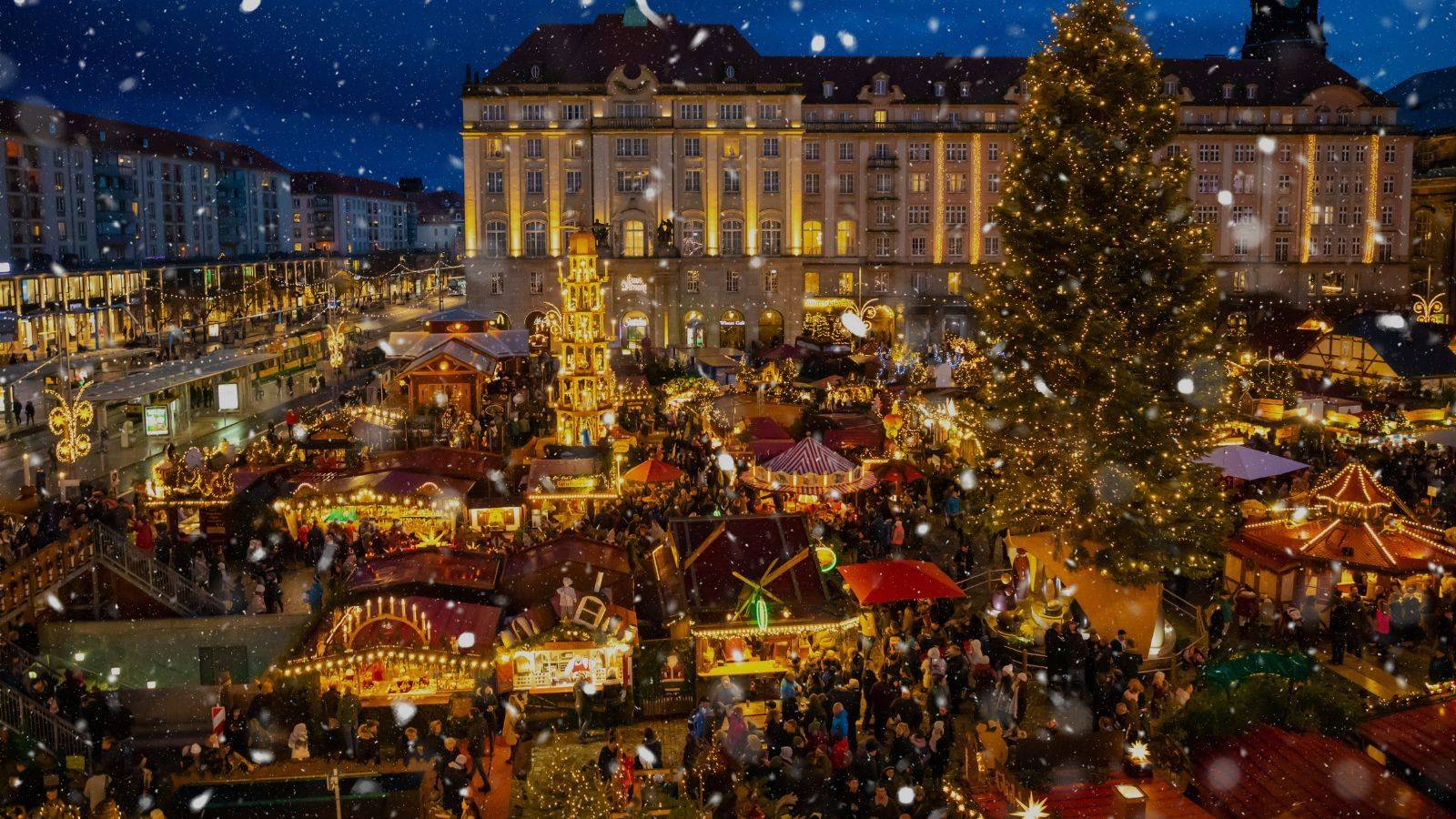 A German Christmas market at night with a lit Christmas tree, wooden stalls and falling snow