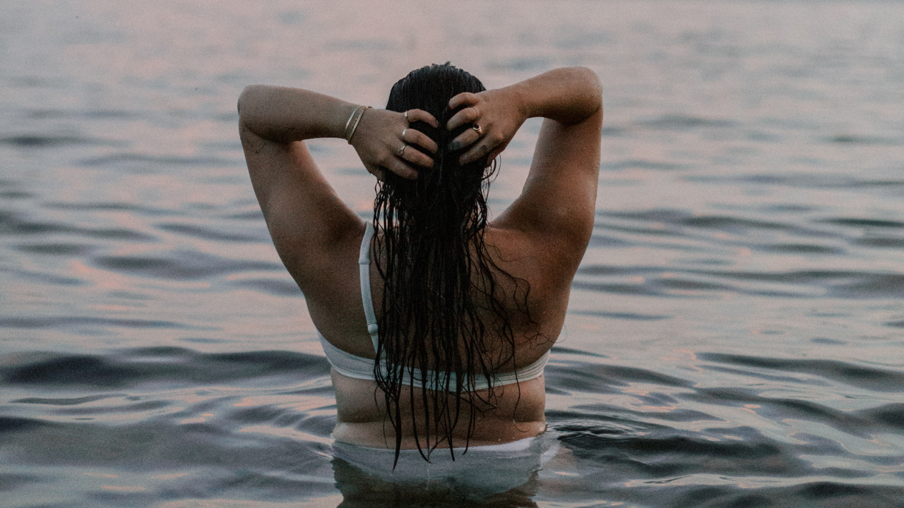 Woman standing in calm water, hands in her hair, facing the horizon — a moment of stillness and intention for female founders