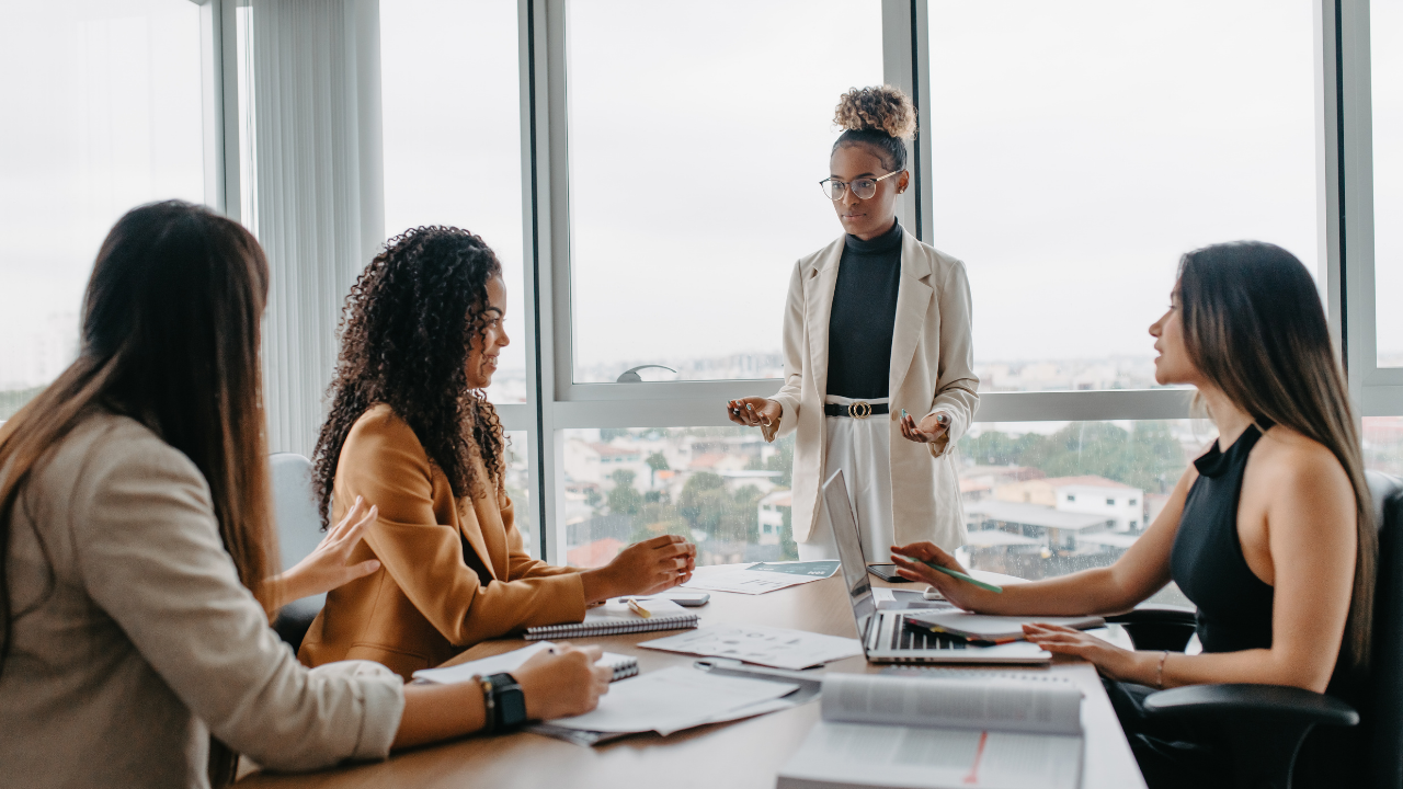 A group of diverse female leaders engaged in a collaborative strategy meeting in a modern, glass-walled boardroom to enhance workplace infrastructure and employee well-being