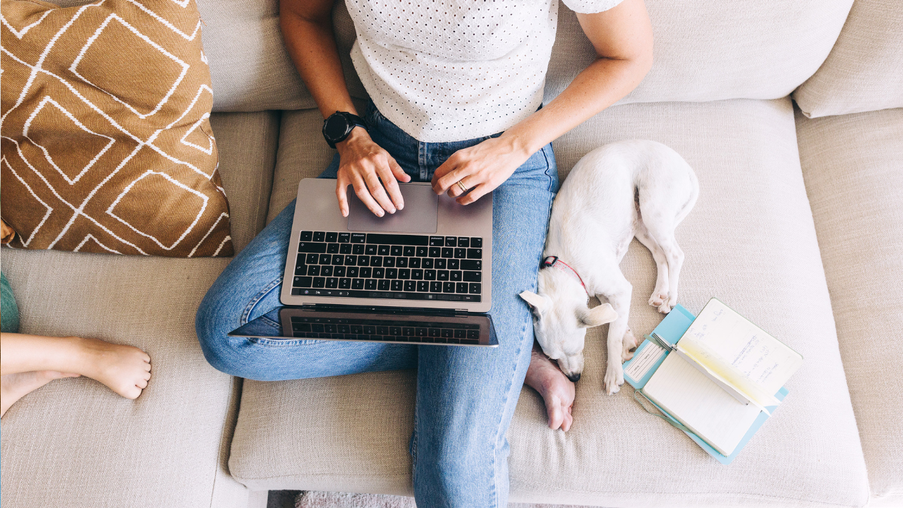 Woman working on a laptop on a sofa with a child and dog, symbolising working from home and work-life imbalance