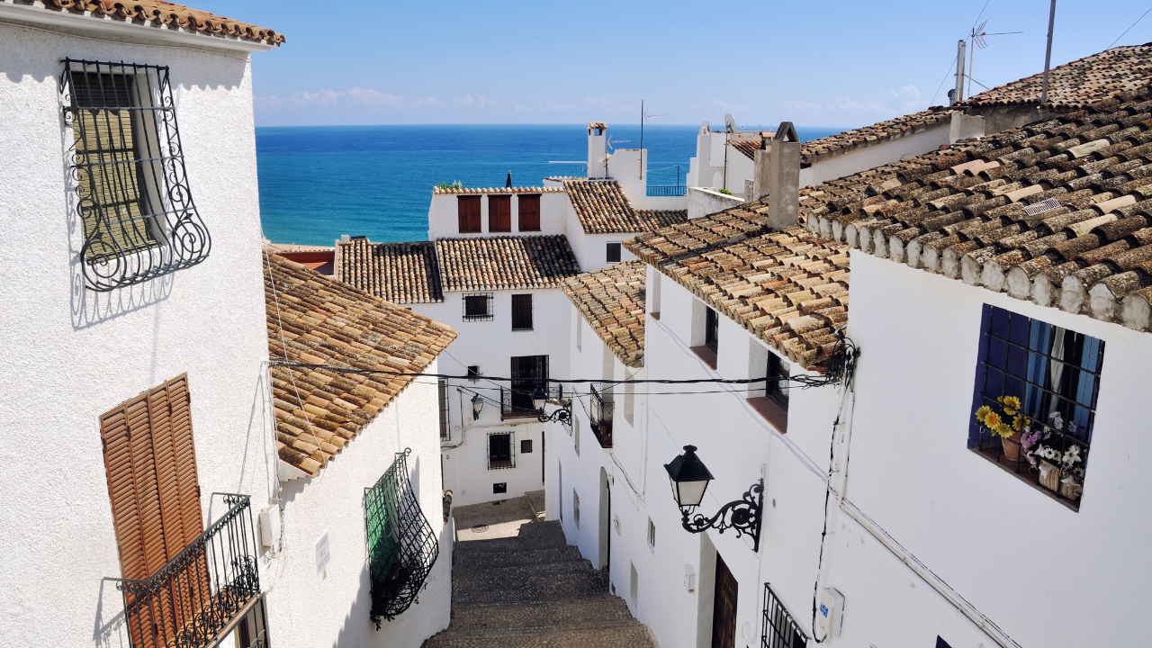 Narrow cobbled street in Altea old town, Spain, lined with white-washed buildings and terracotta roof tiles, with the blue Mediterranean sea visible in the distance