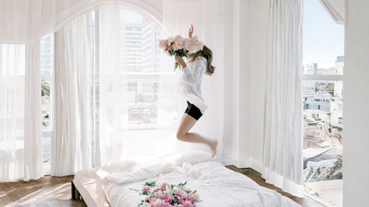 Woman jumping on a bed holding flowers in a bright, sunlit room, capturing a sense of possibility and momentum.