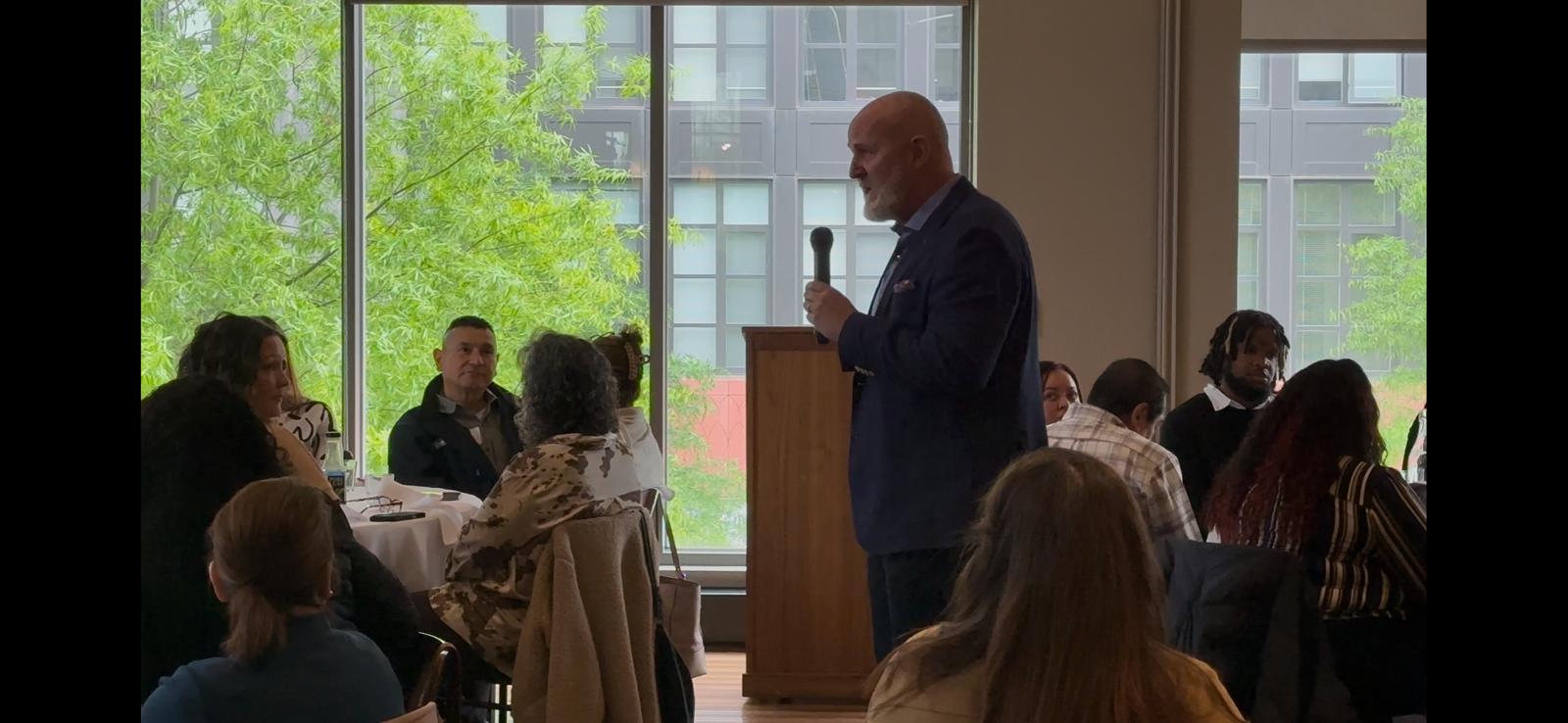 A speaker holding a microphone and addressing a seated audience during a small indoor event or workshop