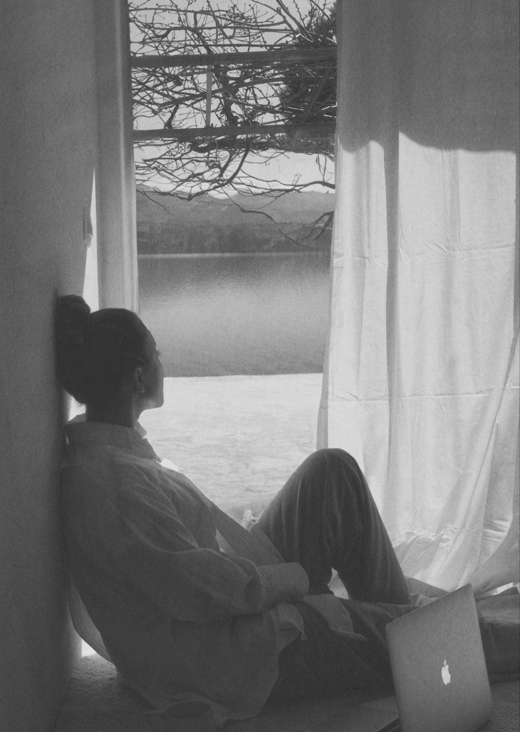 Black-and-white photo of a woman sitting by a window with her laptop closed, gazing out at the water