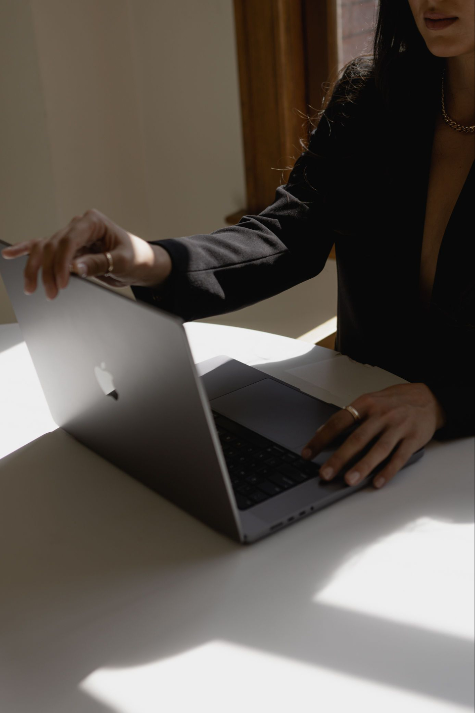 Woman in a black blazer opening her laptop at a sunlit desk
