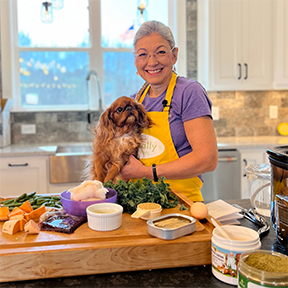 Dr. Judy Morgan holding a Cavalier King Charles Spaniel in a kitchen with fresh pet food ingredients laid out on a cutting board.