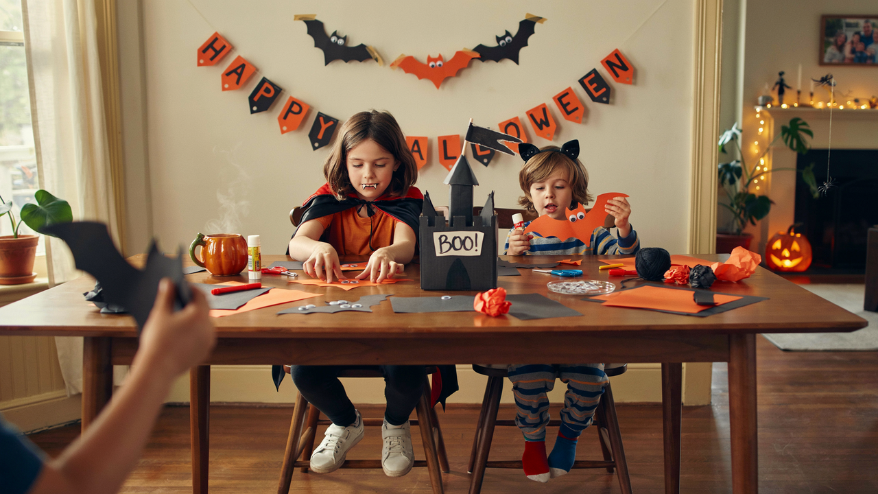 A wide-angle photo of two children in Halloween costumes sitting at a wooden table, crafting with orange paper and a black castle centerpiece. The room is decorated for Halloween with a 'HAPPY HALLOWEEN' banner, bat cutouts, and a lit jack-o'-lantern in the background.