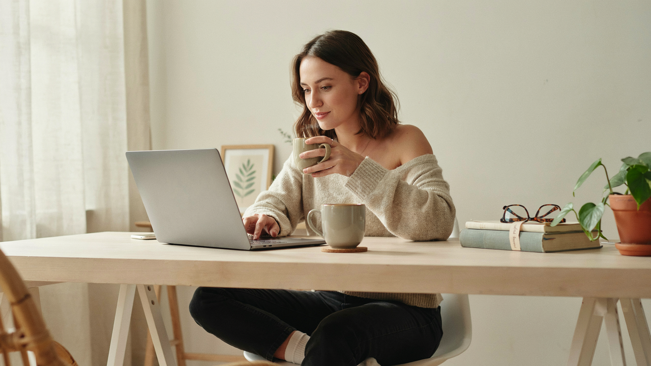 A woman sits at a light wood table in a bright, minimalist room, typing on a silver laptop while holding a green mug. She wears an off-the-shoulder beige sweater and black pants, with a second mug, a stack of books, and a potted plant on the table beside her.