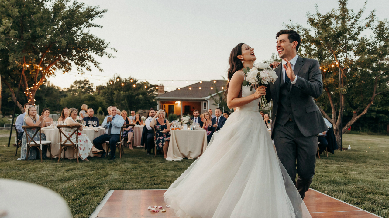 A wide-angle photo of a bride and groom laughing and dancing on a wooden dance floor during an outdoor wedding reception at dusk. The bride wears a white tulle gown and holds a bouquet of white flowers, while the groom wears a dark grey suit. In the background, guests sit at round tables under string lights and trees, with a house visible in the distance.