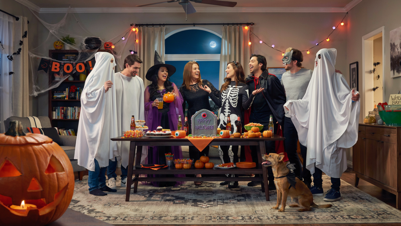 A group of friends dressed in Halloween costumes, including ghosts, a witch, a skeleton, and a vampire, gather around a decorated table in a living room for a party. The scene is filled with festive decorations like spiderwebs, string lights, and a large carved jack-o'-lantern in the foreground. A cozy, well-lit living room decorated for Halloween. The walls are a neutral beige, and the ceiling features a dark wood ceiling fan with five blades. A large arched window in the center background shows a dark night sky with a full moon. The room is decorated with white spiderwebs draped across the ceiling and walls, orange and purple string lights, and various Halloween-themed decorations including a 'BOO' banner and a jack-o'-lantern. A dark wood sideboard stands on the right, and a bookshelf is visible on the left. The floor is covered by a large, patterned area rug in shades of beige and brown.