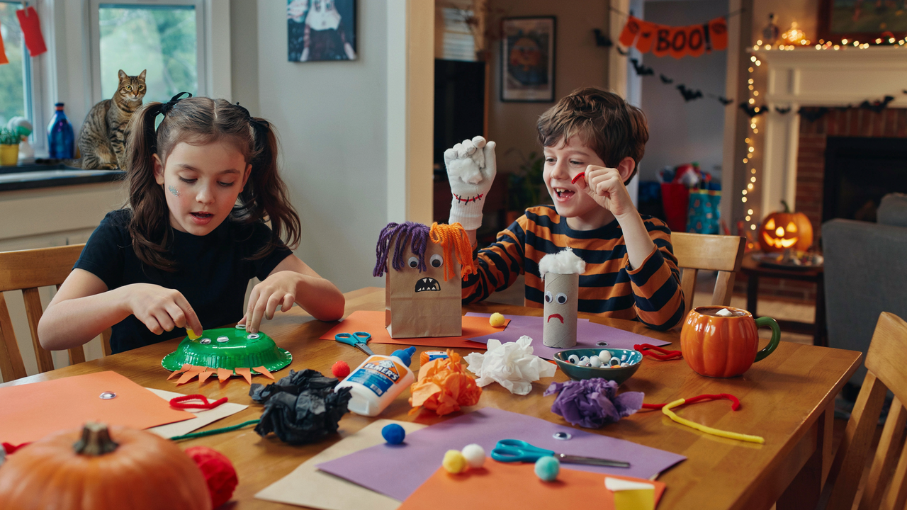 A wide-angle photo of two children sitting at a wooden dining table, engaged in Halloween-themed arts and crafts with paper bags, pom-poms, and glue. A tabby cat sits on a kitchen counter in the background, and the room is decorated with orange paper banners and a lit jack-o'-lantern.